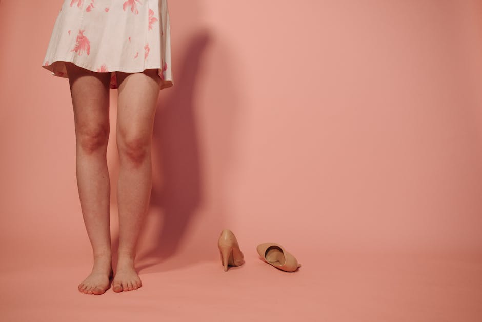 A woman's legs in a floral skirt with nude high heels on a peach background, barefooted.